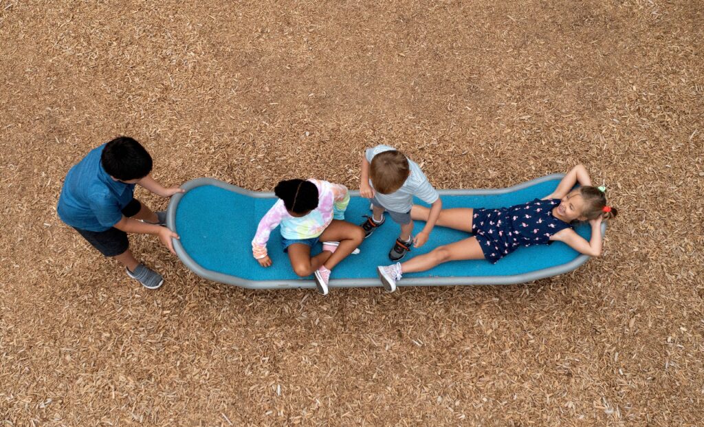 An aerial view of four children playing together on a blue, surfboard-shaped inclusive rocker in a playground with woodchip flooring. One child pushes from the end while three others sit, stand, and lie down on the wide, accessible platform.
