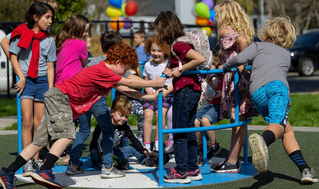A group of children playing together on a blue and white inclusive roundabout at a playground on a sunny day.