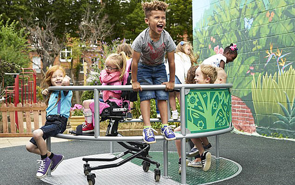 A group of diverse young children laughing and playing together on an inclusive playground roundabout. One child uses a supportive seating device integrated into the equipment.