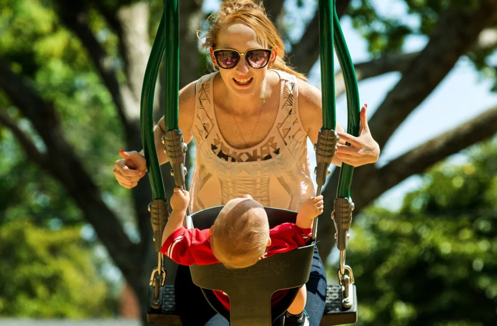 A woman wearing sunglasses and a young child enjoying a ride together on a green inclusive Buddy Swing.